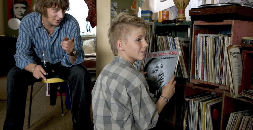 Still-billede fra filmen "Drømmen" (2006). To personer i en stue, hvor den ene dreng holder et vinylalbum med Martin Luther King, mens den anden sidder på en stol.