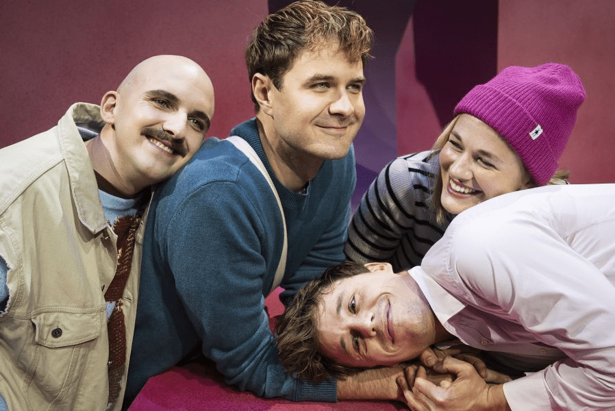 Fire skuespillere poserer sammen på en farverig scene, mens de smiler og interagerer med hinanden.